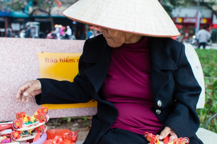 Old woman sells small wooden toys in Hanoi park.